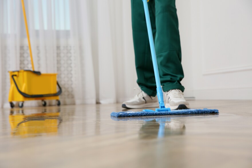 A person seen from the knees down and wearing what look like green scrubs is pushing a blue mop across the floor. The floor is smoothy an reflective where it has just been cleaned, and a yellow bucket, also reflecting against the shiny floor, is in the background.
