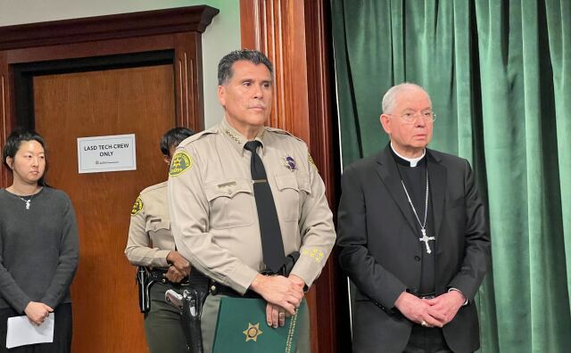A man with medium-tone skin wears a tan sheriff's uniform while standing  with his arms crossed. Next to hom is a man in a priest's collar and wearing a cross in all black. 