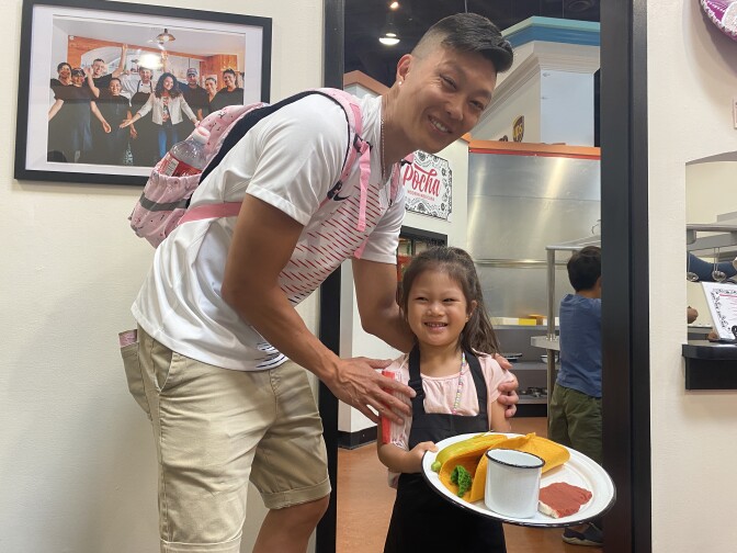 An Asian man in a white shirt stands smiling with his arms on the shoulders of a 4-year-old Asian girl, who is also smiling. She wears a blue apron and has a plate full of fake food in her hands.