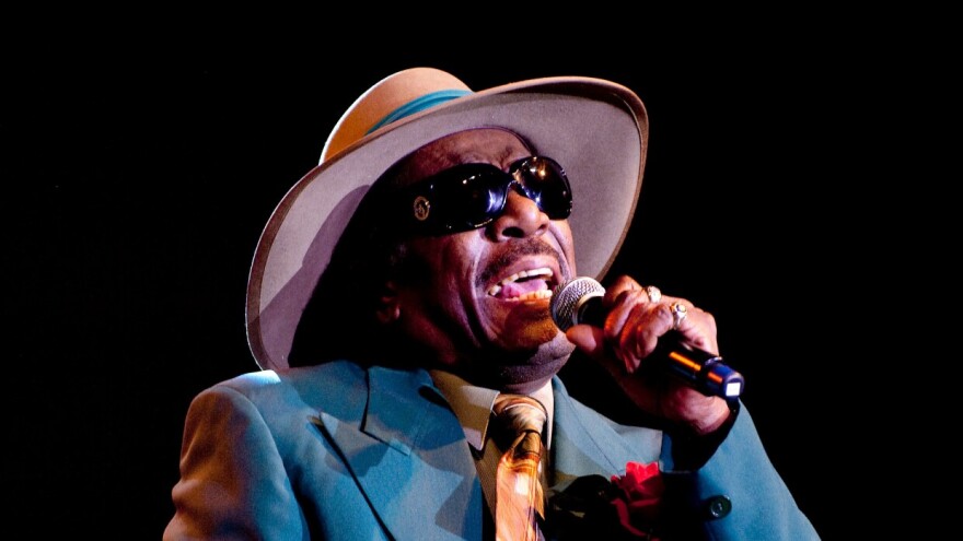 A Black man in a blue suit wears a wide-brim gat as he sings into a mic. He has sunglasses on.