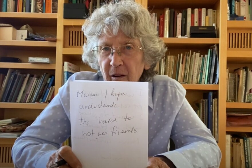 An older woman with light skin tone sits in front of a full bookcase. She holds up a piece of paper to the camera. The paper has a handwritten note that says "Mama/Papa understands. It's hard to not see friends."