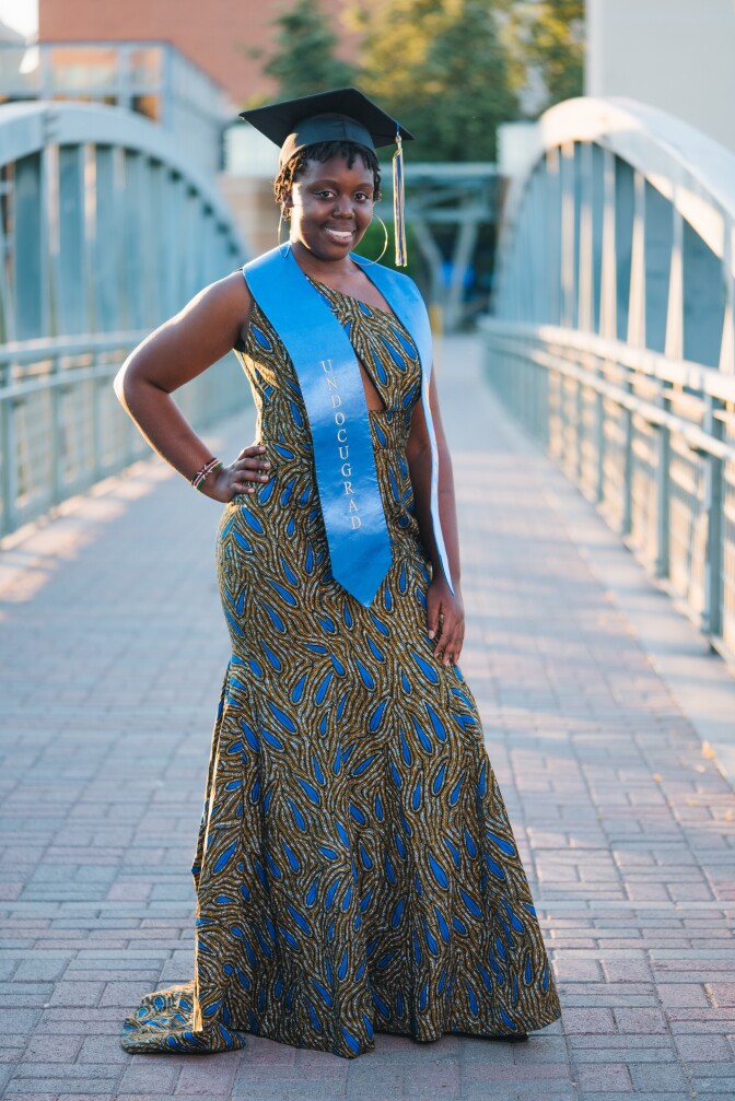 A Black woman with short hair wearing a graduation cap and a blue sash that reads "Undocugrad" stands in front of a bridge, hand on hip. She's wearing a long blue and yellow patterned dress. 