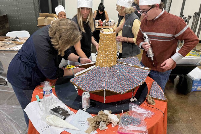 A group of people wearing chef hats are working to build a gingerbread structure