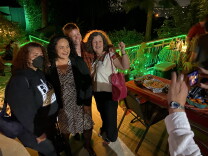 Four women pose and smile for a photograph on an outdoor patio at nighttime. On the left of the photo, a woman dressed in a sweatshirt that says "Black Lives Matter At School" wears a facemask. One brown-skinned woman with curly dark-brown hair in the middle of the group — school board candidate Rocio Rivas — smiles in a black-and-white dress. A woman on the right side of the photo raises a fist and smiles.