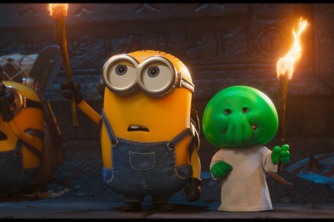 A still of an animated movie, a pill shaped yellow creature wearing overalls holds up a torch, two more creatures like him are in the background. A green smaller creature holding a torch is also present.
