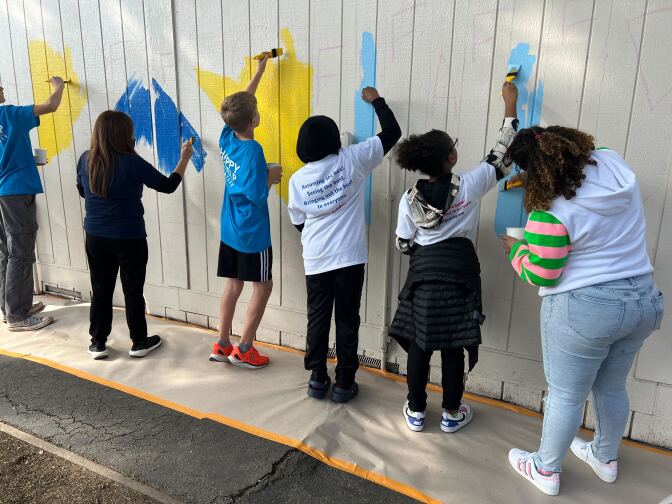 Six children, all with their backs to the camera, apply blue, yellow and light blue paint to a fence as part of a mural project