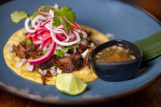 A beef taco on a blue plate with garnishes and au jus on the side. 
