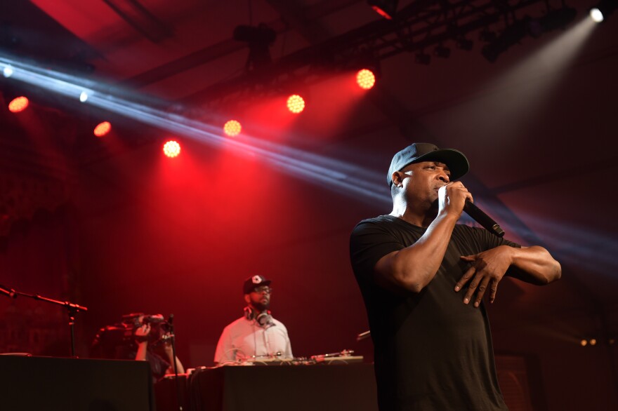 Rapper Chuck D, A middle-aged Black man, stands on a stage in front of a DJ and red lighting, holding a microphone. He wears a black t-shirt and a black hat.