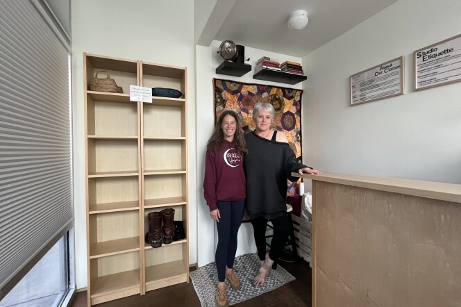 Two light-skin toned women stand side by side behind a desk.  One woman to the left is wearing a burgundy hoodie, black leggings and brown shoes and she has medium to long brown wavy hair. The other woman to the right is wearing a grey sweater tunic, with black leggings and she has short grey hair. To their left is a shelf filled with shoes, a basket and a pillow. 