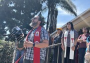 A man with a beard wearing a red stole speaks into microphones at a news conference setting.