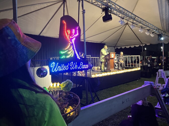 A neon sign that reads "United We Dance" is hung next to the stage where a man stands at a podium. 