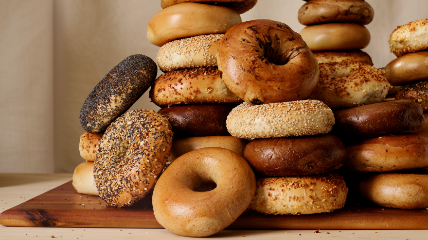 A pile of brown shiny bagels, some with poppy seeds, some with sesame seeds, some with everything, some plain: They all sit on a wooden cutting board placed atop a counter.