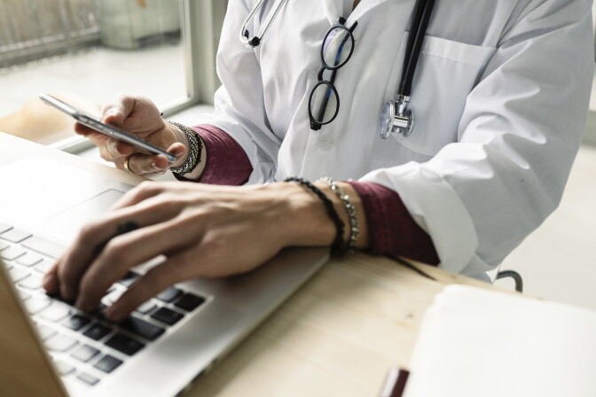 A person wearing a white shirt with the sleeves rolled up and a burgundy shirt underneath. Eyeglasses hang from their shirt and a stethescope is around their neck. In one hand they are holding a cellphone, the other rests on the keyboard of a silver laptop.
