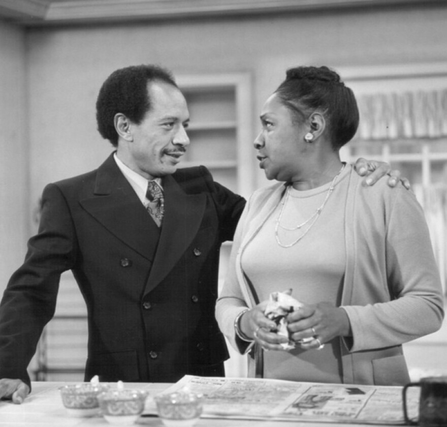 A Black man in a double-breasted suit has his arm around the shoulder of a Black woman in a sweater set. They're standing at a kitchen counter.