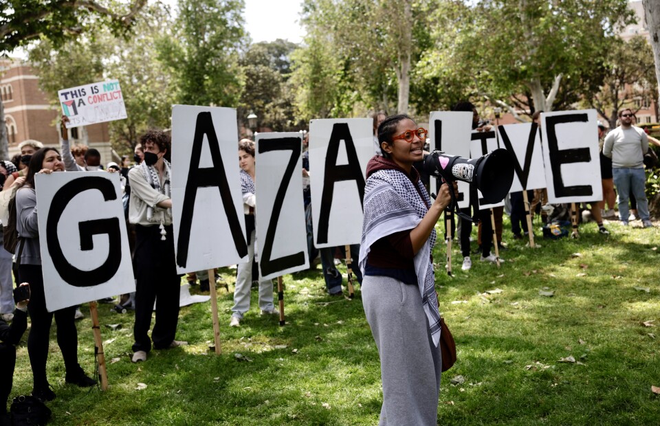 A person with medium skin tone and red framed glasses wears a Keffiyeh around their shoulders while speaking into a megaphone. Behind them are people holding large letters that spell out "Gaza Live"