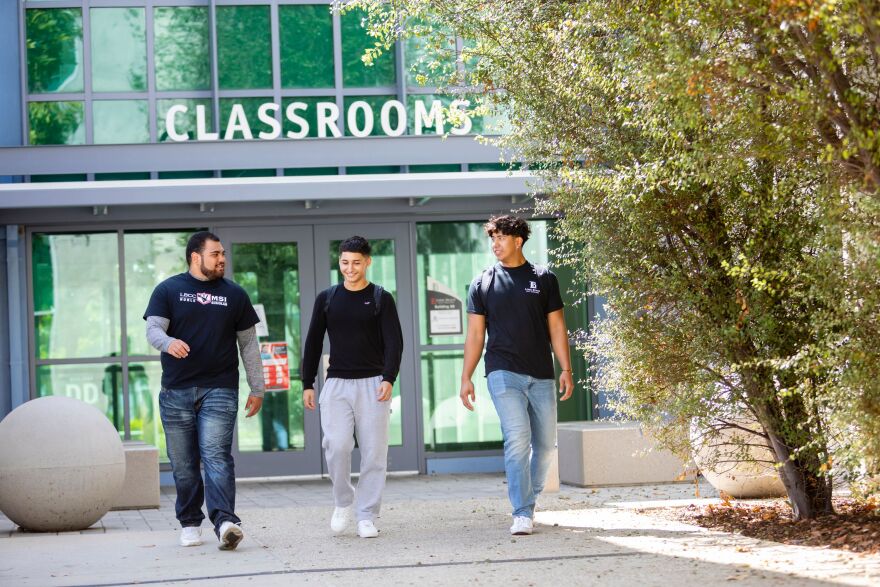Three students, all wearing black t-shirts, jeans, and sneakers, walk by a campus building with a sign above the entry that says 'Classrooms'