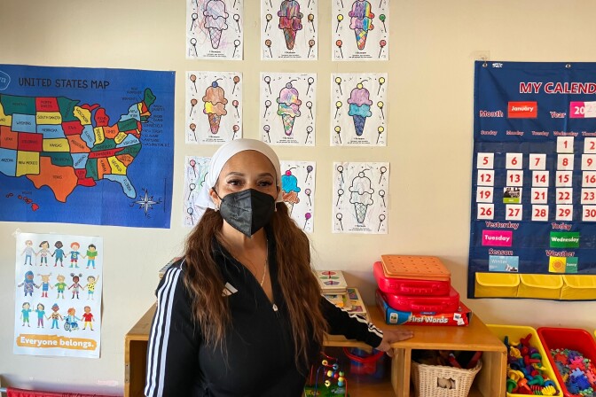 A woman with medium skin tone wears a black facemask and stands for a portrait in front of a wall decorated with colorful signs and posters.