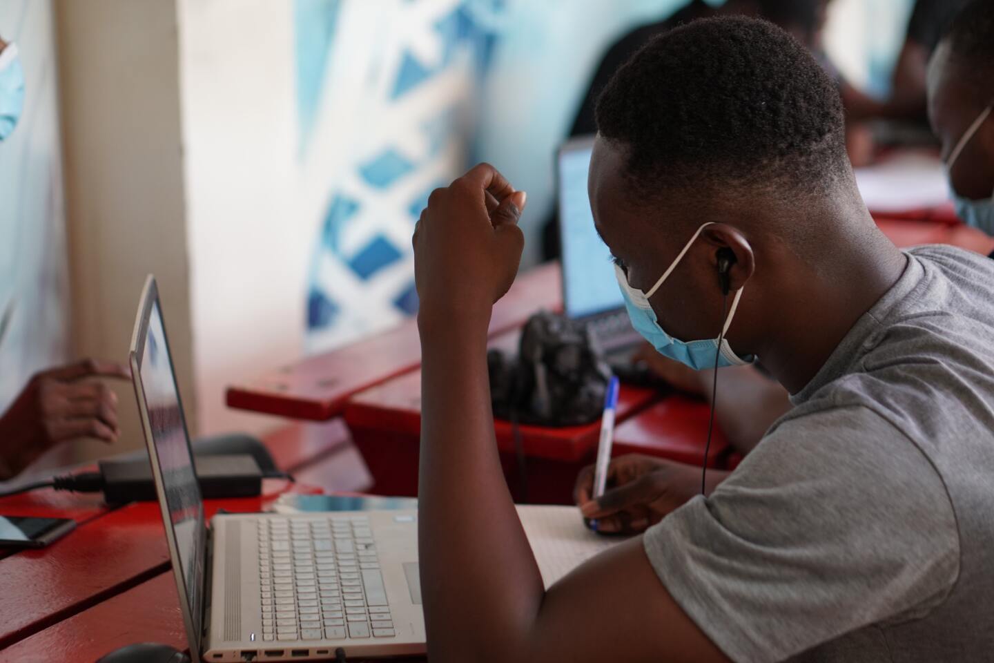 A side profile view of a man wearing a face mask as he works on his laptop at an indoor cafe.