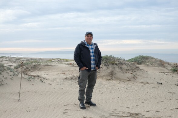 A middle aged man with light tan skin wears a black jacket, blue and white plaid shirt and dark pants, stands on the beach with small dunes covered with low-lying plants behind him. The ocean is hazy beyond. 