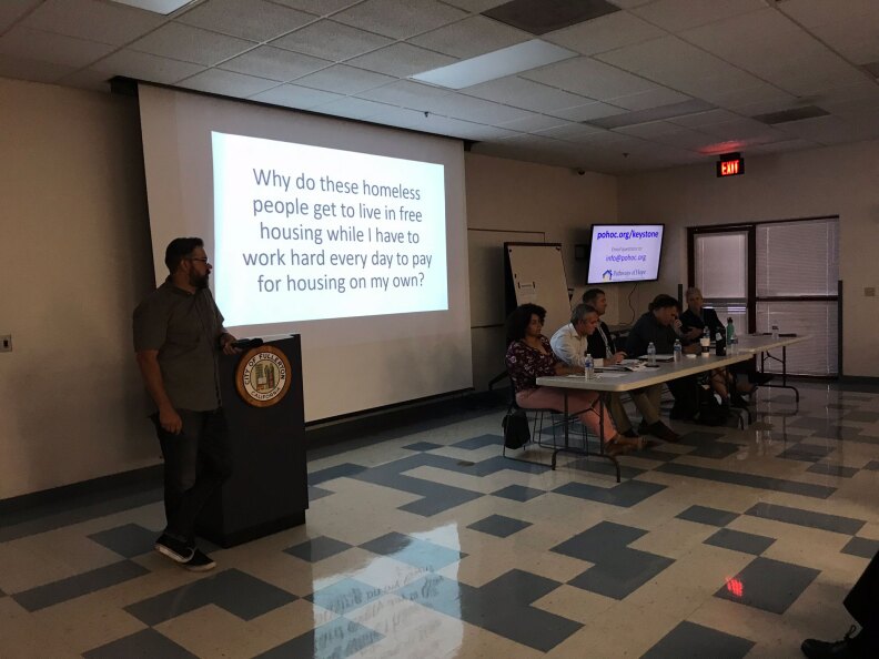 A man stands by a projector screen on a wall that says, "Why do these homeless people get to live in free housing while I have to work hard every day to pay for housing on my own?"