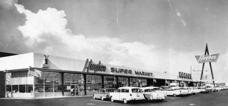 A grocery store anchors a strip mall with big windows. Vintage cars and an American flag are visible as well as multiples rights for Hughes Super Market