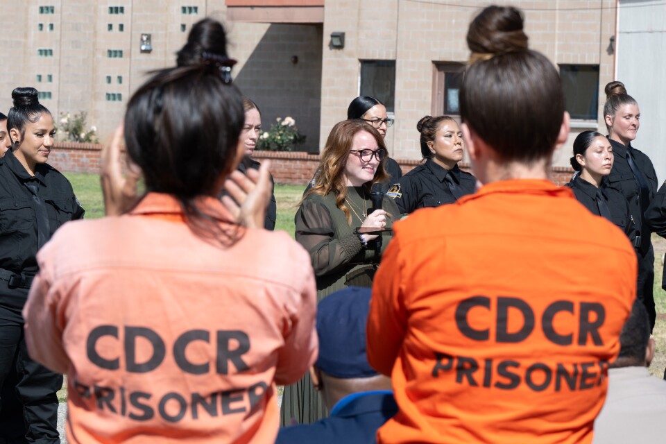 Two women wearing orange jumpsuits that say "CDCR prisoner" look on as a woman speaks into a microphone.
