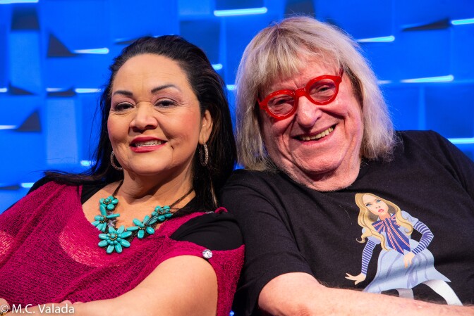 Josefina López, a Latina woman wearing a read dress and turquoise necklace and Bruce Vilanch, a white man with long blond hair wearing bright red glasses, smile at camera