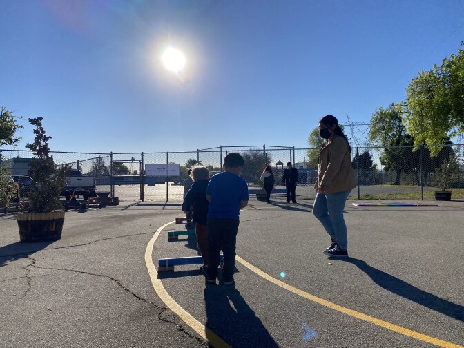 Young children play with tiny hurdles placed between striped lines outlining a racecourse on a blacktop surface as a teacher looks on.