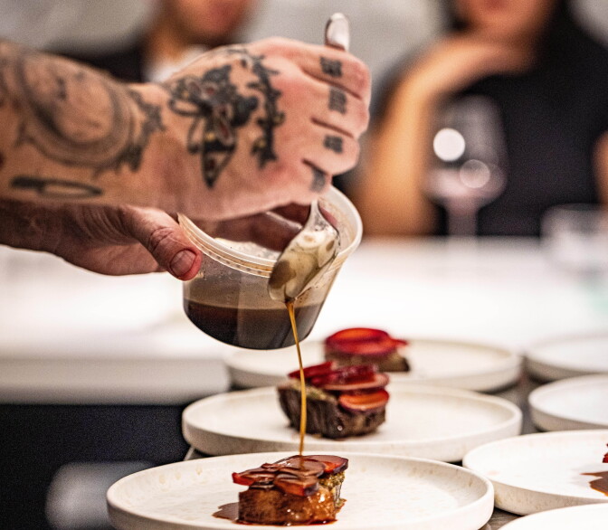 A pair of light skinned hands with several tattoos spoons a brown liquid over a series of white plates which each hold a piece of beef.