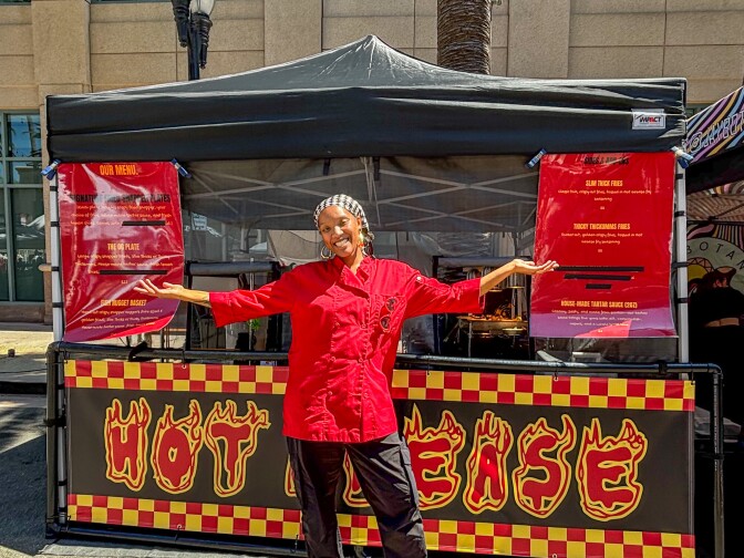 A woman with dark skin smiling in a bold red chef’s jacket and patterned headscarf stands proudly in front of her “Hot Grease” stall,  with her arms outstretched, framed by sizzling menu boards and the hum of the street market behind her.