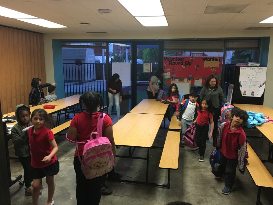 A number of young students with varying skin tones stand around a small room filled with long picnic tables.