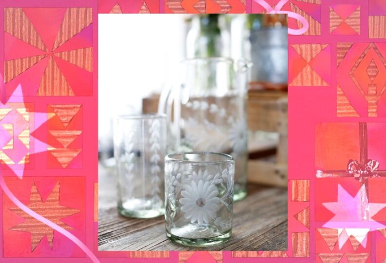 A rustic wooden tabletop is set with glasses of varying sizes, and a drinking pitcher. All the decoratively etched in some way, one glass has ornage flowers on it, another class has etched hearts, while the pitcher is etched with both patterns. The photograph is set against a decorative pink background accented with stars, gift boxes and strands of ribbon.