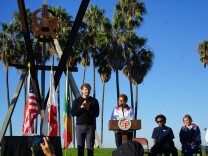 A woman stands behind a podium and a man motions beside her. Several other people stand behind them with palm trees in the distance. 