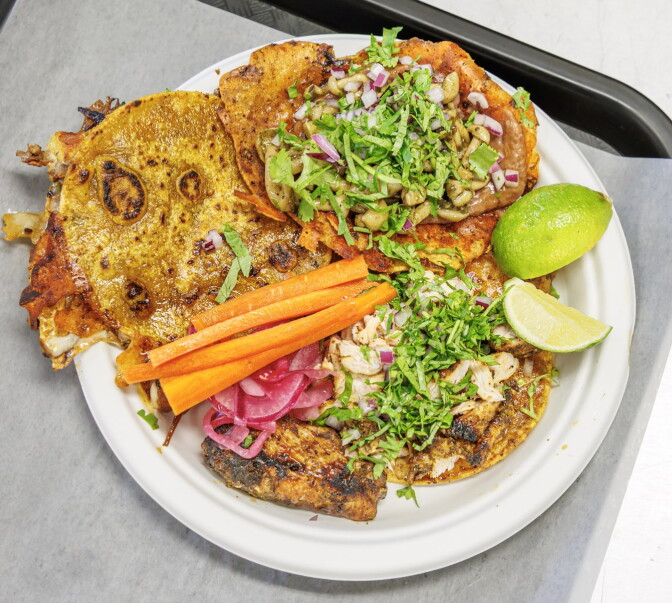 Overhead view of a white paper plate holding three tacos on crisp, griddled tortillas: one folded with melted cheese along the edges, one topped with mushrooms, refried beans, red onion, and cilantro, and one topped with grilled chicken, onion, and cilantro. Carrot sticks, pickled red onions, and lime wedges sit alongside.