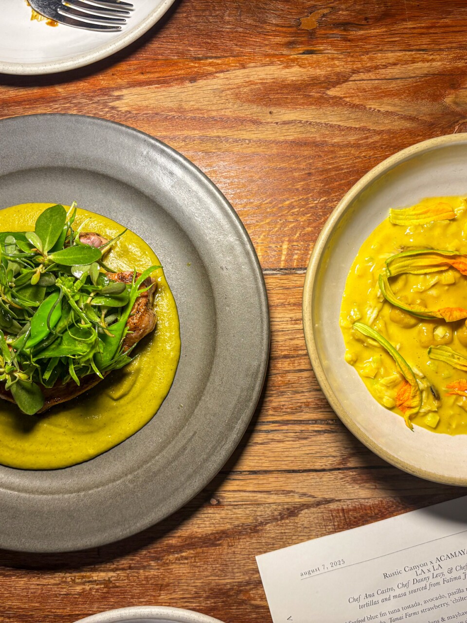 Two plates on a wooden table show chochoyotes with squash blossoms in golden-green sauce on top, and roast chicken with pepián sauce topped with fresh greens and edible flowers on bottom.