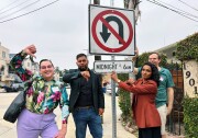 A wide shot of four people with medium and light skin tones standing near a street sign outside, looking toward the camer and showing a thumbs down hand gesture. The sign is a red and black no u-turn sign with a timeframe below that reads Midnight to 6 a.m.
