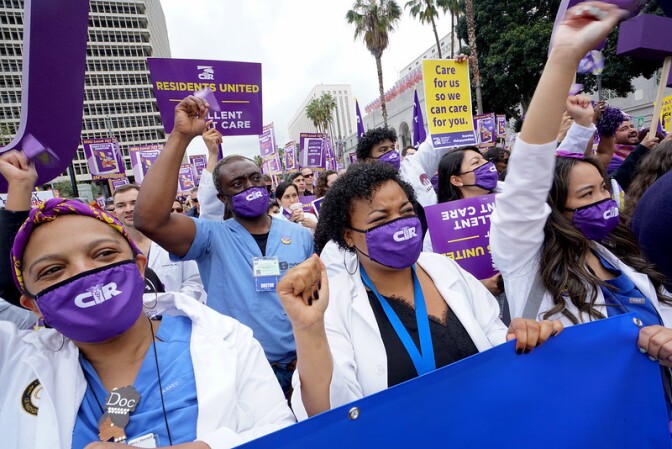A group of people wearing purple masks and raise their fists in the air. There are hundreds of people around them with protests signs saying,"Residents united" and  "Care for us so we can care for you."