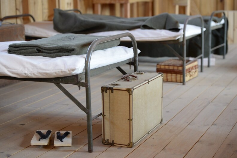 Interior of a Japanese internment camp, with cots, blankets, suitcases and a pair of slippers