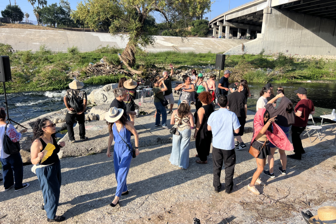A group of people dancing in the concrete bed of the Los Angeles River is depicted. Speakers are seen on either side of the picture and a large tree is seen in the background.
