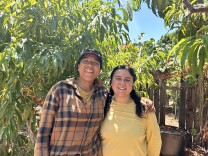 Two women are smiling at the camera with their arms around each other. The woman on the left is wearing an orange long-sleeve flannel and a gray baseball cap, while the woman on the right has her black hair pulled back in a ponytail and is wearing a pale-yellow shirt. They're both standing in an outdoor farm space filled with leaves and trees on a sunny, clear morning.