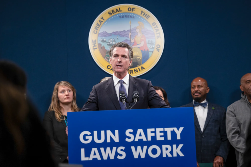 Governor Gavin Newsom, a light-skinned man with salt-and-pepper hair wearing a charcoal gray suit and blue tie, stands at a podium labeled GUN SAFETY LAWS WORK in white lettering on a blue background. The California state seal is posted behind him. Four people stand behind him a the podium.