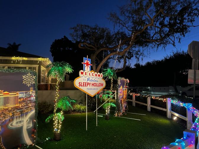 An elaborate holiday light display in the front yard of a home. Lights are wrapped around the white picket fence and fake palm trees, with a large side that reads "Welcome to Fabulous Sleepy Hollow Torrance" sticking out of the green grass in the center. 