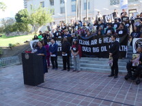 A person speaks at a podium at the steps of L.A. City Hall as a crowd behind her holds banners. One reads"Clean Air & Health Over Big Oil Wealth."