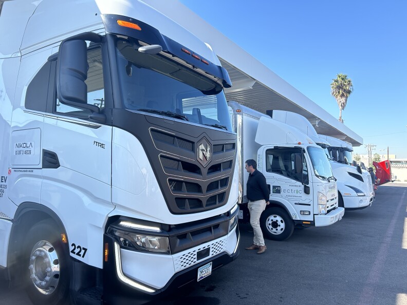 Large white big rig electric trucks lined up in a parking lot under a blue sky. 