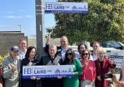 A group of people, smiling and holding thumbs up, huddle around a sign that reads "Honorary Laird Lane."