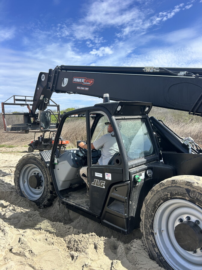 A person operates a heavy-duty sand-handling machine with its forklift extended, parked on a beach with a marsh landscape behind.