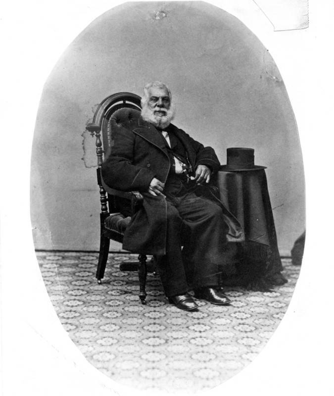 A black and white photos shows a Black and Indigenous Mexican man with a fluffy white beard and white hair wears a black coat, black pants and black shoes, and sits in a black chair. A table draped with a black tablecloth is to his right, his arm is resting on it. On top of the table is a tall black hat. The floor is tiled with a geometric pattern.