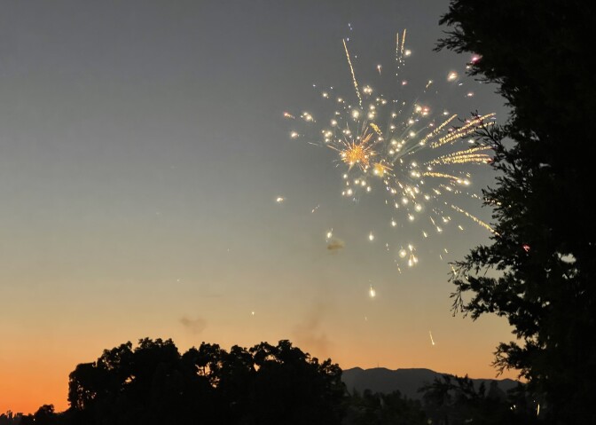A firework sparkles silver, yellow and orange at sunset, with trees and hills framing the image.