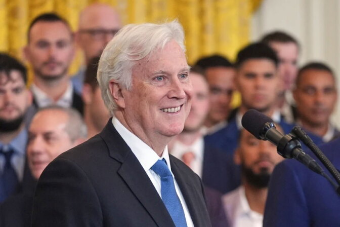 A man with white hair and a light skin tone smiles in a dark suit and blue tie, with a microphone in the foreground and a group of people in suits behind them.