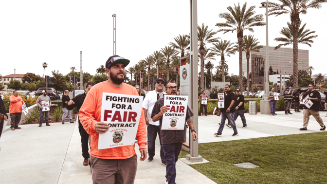 Men walk in front of county buildings holding signs that say, "Fighting for a fair contract." 
